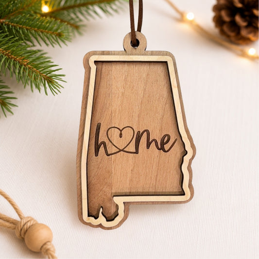 Layered Alabama wood ornament featuring engraved “home” script and faux leather hanger on a soft neutral holiday background with greenery and warm lights.