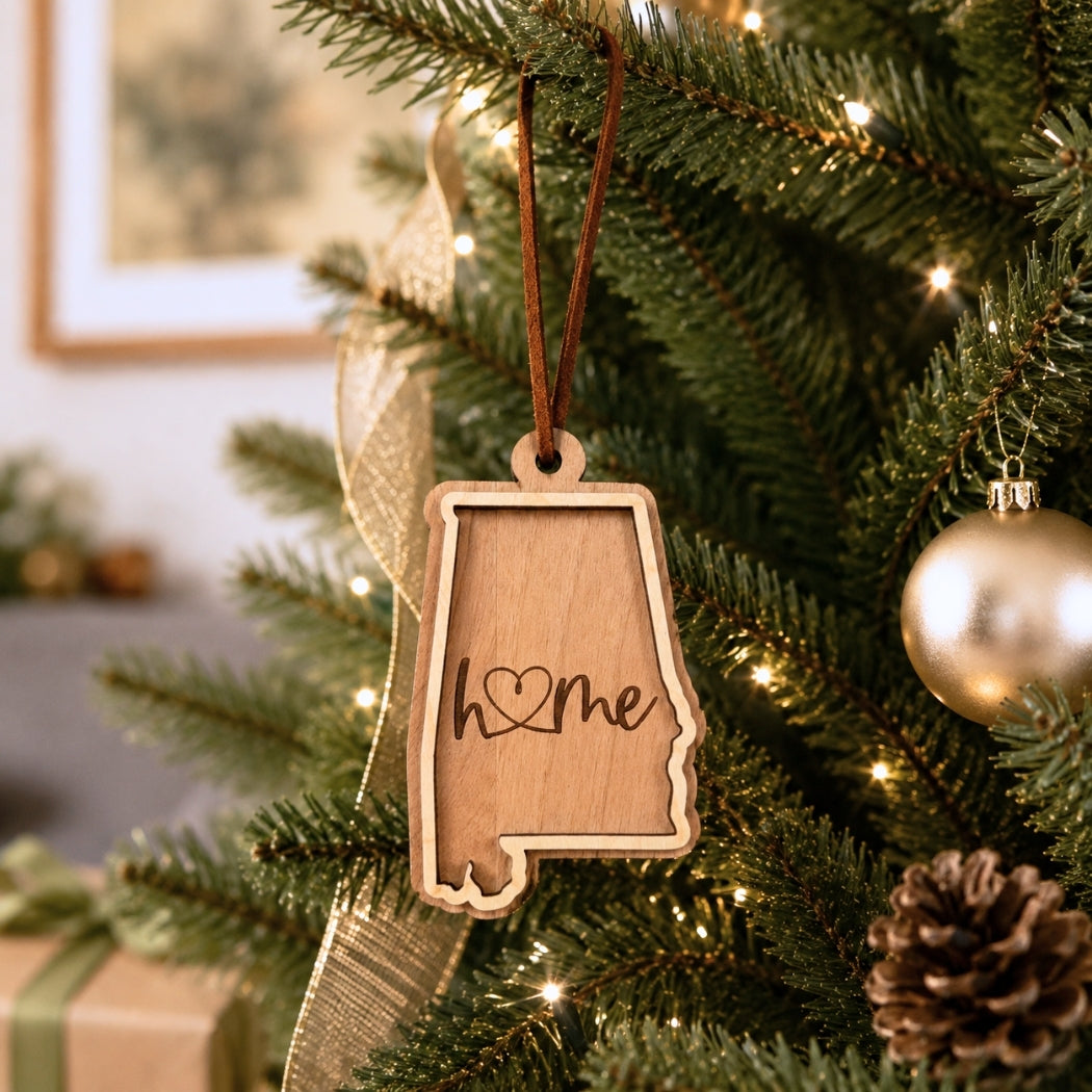 Layered Alabama “home” ornament hanging on a decorated Christmas tree with warm white lights, pinecones, and gold ornaments.
