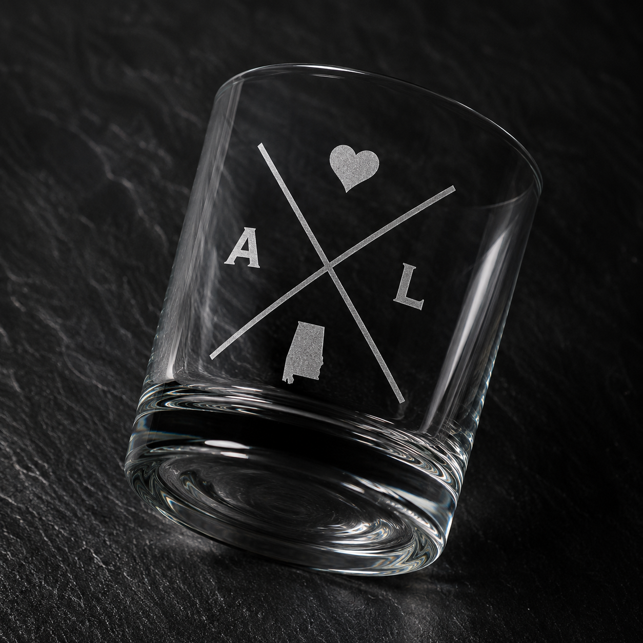 Angled engraved Alabama whiskey glass featuring frosted Alabama silhouette and modern crossed-letter design on a dark textured surface.