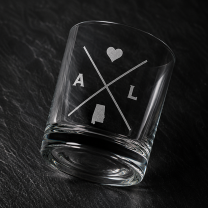 Angled engraved Alabama whiskey glass featuring frosted Alabama silhouette and modern crossed-letter design on a dark textured surface.