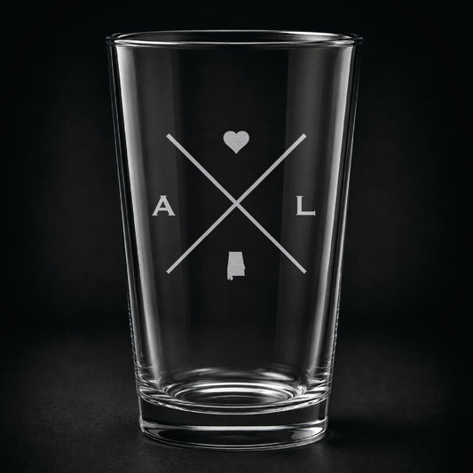Engraved Alabama pint glass featuring a minimalist crossed-line design with heart detail and Alabama silhouette on a dark background.