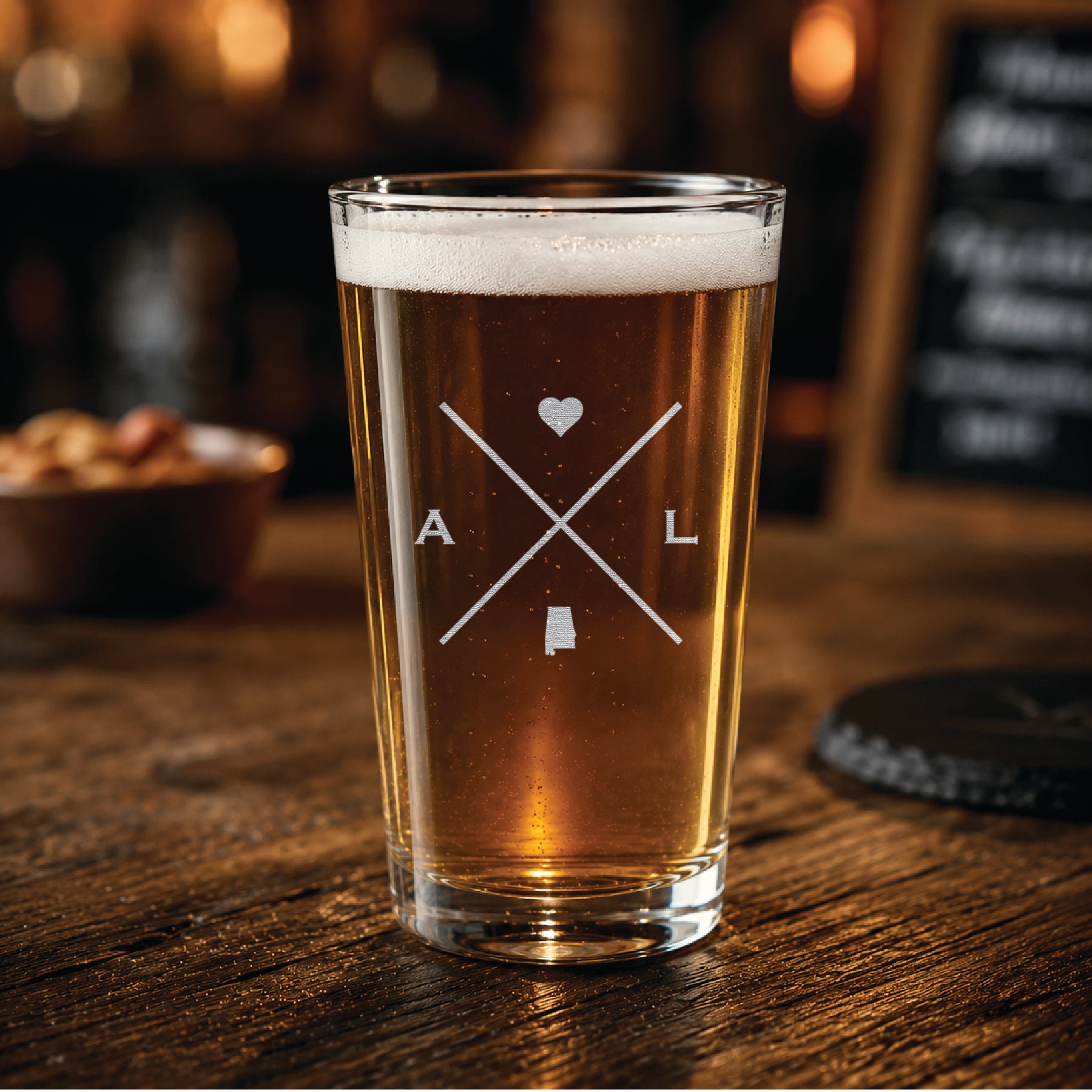 Engraved Alabama pint glass filled with amber beer in a warm pub-style setting, featuring a minimalist crossed-line Alabama design with heart detail.