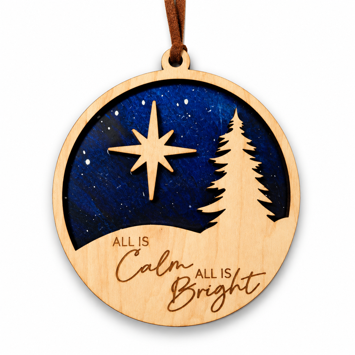 A round wooden Christmas ornament featuring a laser-cut night sky design with a star, pine tree, and the words 'ALL IS Calm ALL IS Bright'. The ornament has a natural light wood border and a dark blue center, hanging from a brown leather loop.