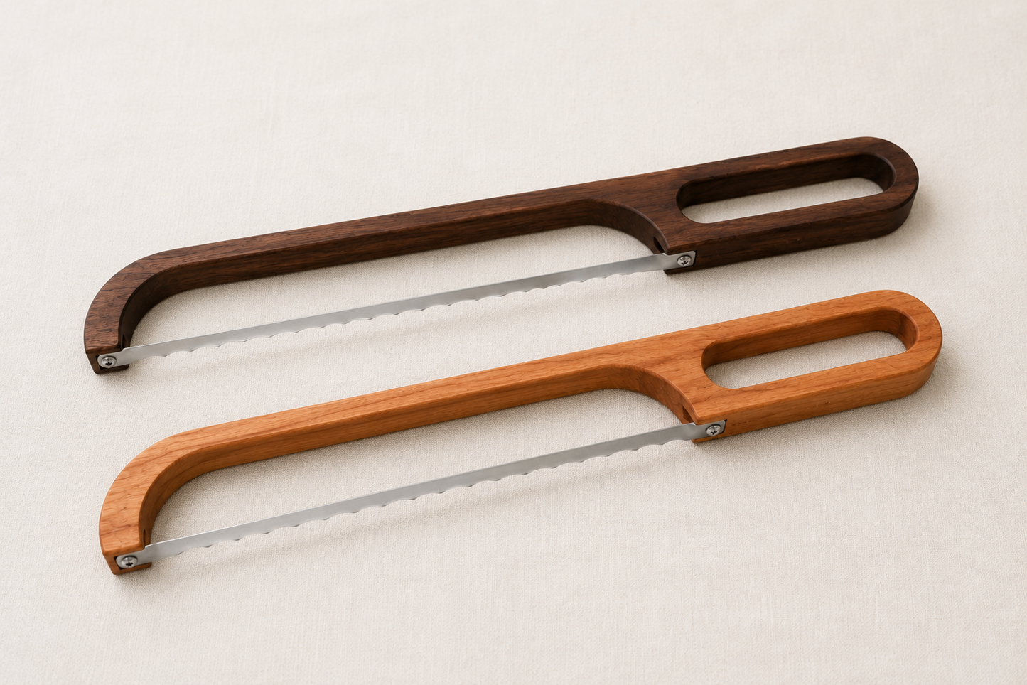 Minimal studio comparison image of walnut and cherry bread bow knives displayed side by side on a neutral fabric background.