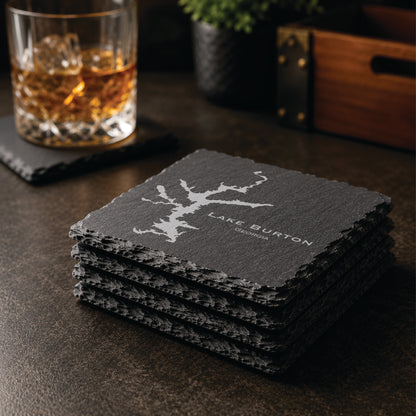 Stack of engraved Lake Burton slate coasters in a warm bar-style setting with whiskey glass nearby, featuring a detailed lake map design and Georgia text.