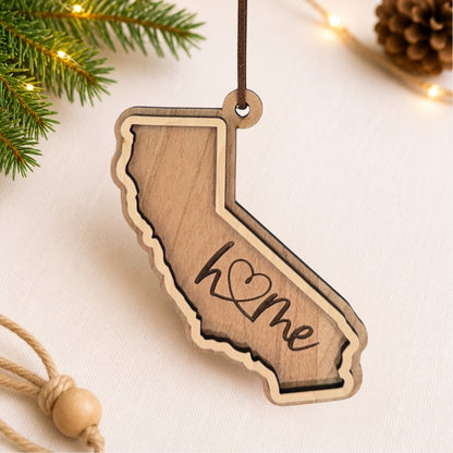 Layered California wood ornament featuring engraved “home” script and faux leather hanger on a soft neutral holiday background with greenery and warm lights.