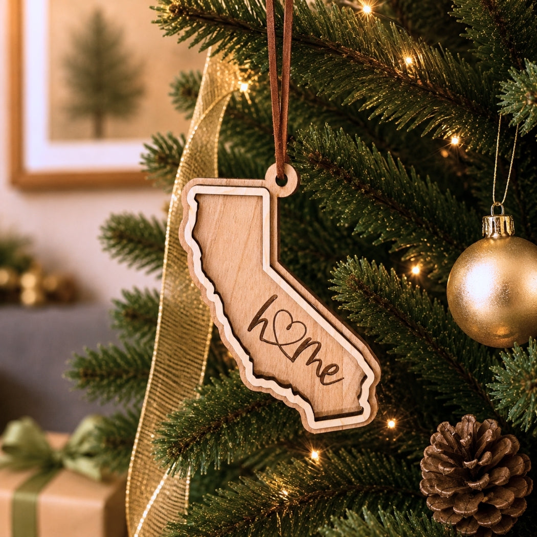 Layered California “home” ornament hanging on a decorated Christmas tree with warm white lights, pinecones, and gold ornaments.