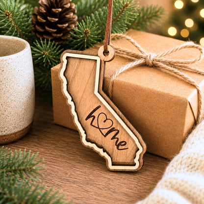 Layered California wood ornament with engraved “home” script displayed beside wrapped holiday gifts, greenery, and warm seasonal décor.