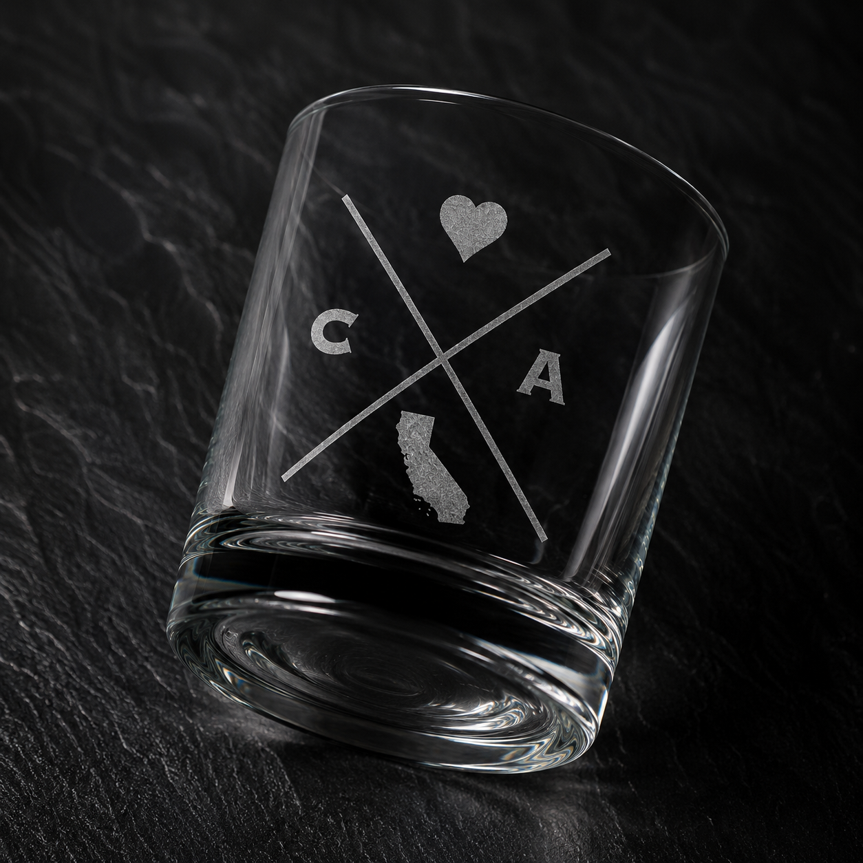 Angled engraved California whiskey glass featuring frosted California silhouette and modern crossed-letter design on a dark textured surface.