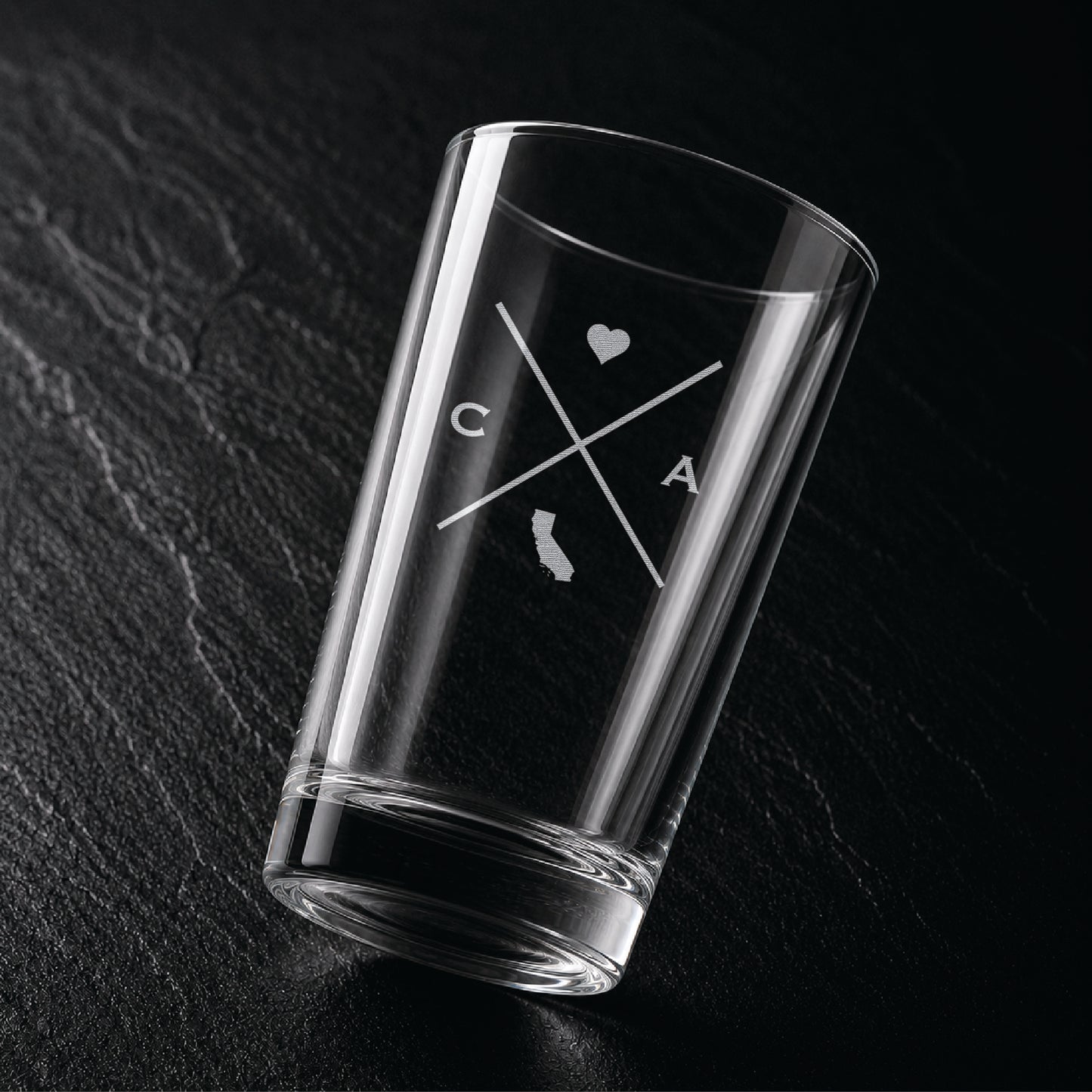 Angled view of engraved California pint glass with modern crossed-line state design and frosted laser engraving on dark textured surface.