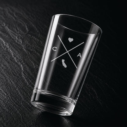 Angled view of engraved California pint glass with modern crossed-line state design and frosted laser engraving on dark textured surface.