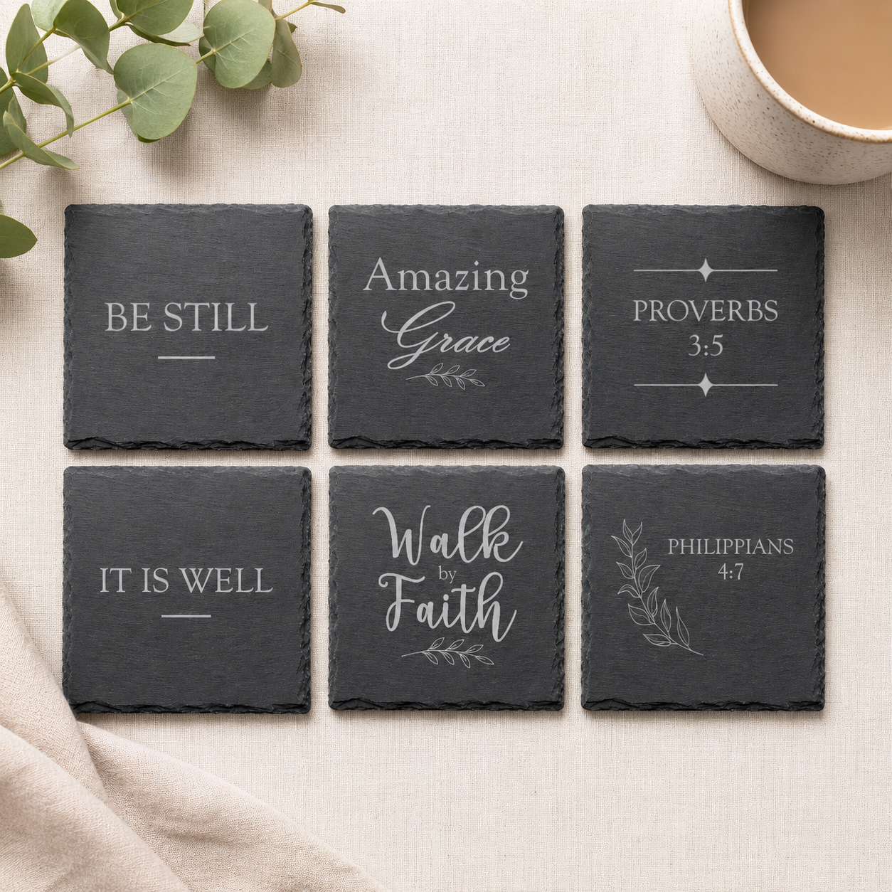 Set of six engraved Christian slate coasters featuring inspirational faith sayings and scripture references displayed on a neutral fabric background with eucalyptus greenery.