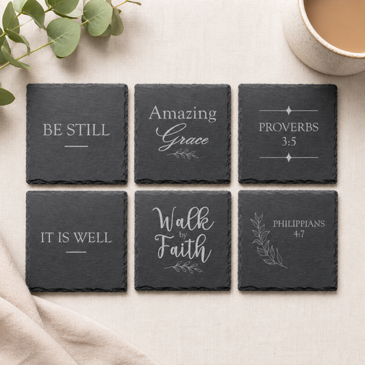 Set of six engraved Christian slate coasters featuring inspirational faith sayings and scripture references displayed on a neutral fabric background with eucalyptus greenery.