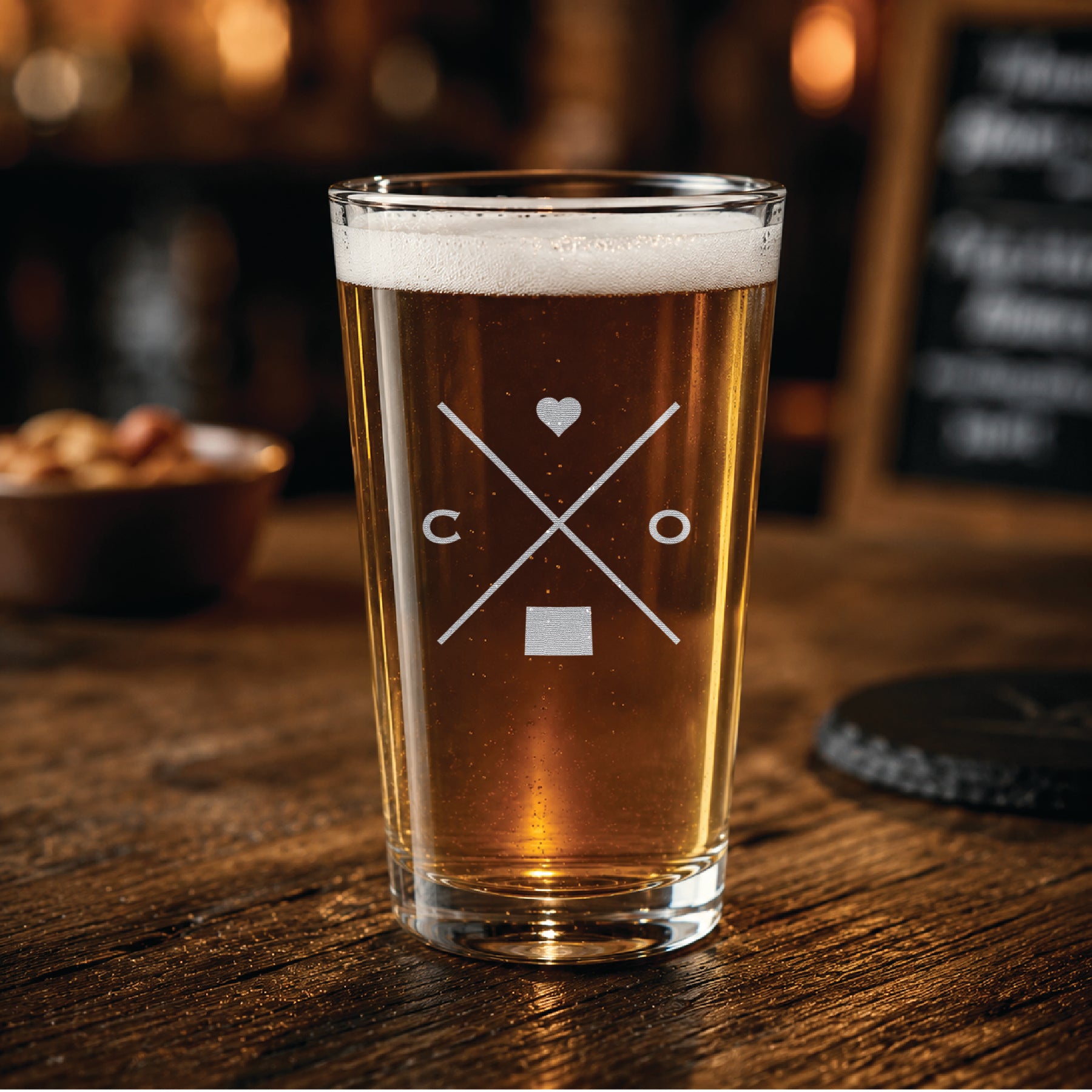 Engraved Colorado pint glass filled with amber beer in a warm pub-style setting, featuring a minimalist crossed-line Colorado design with heart detail.