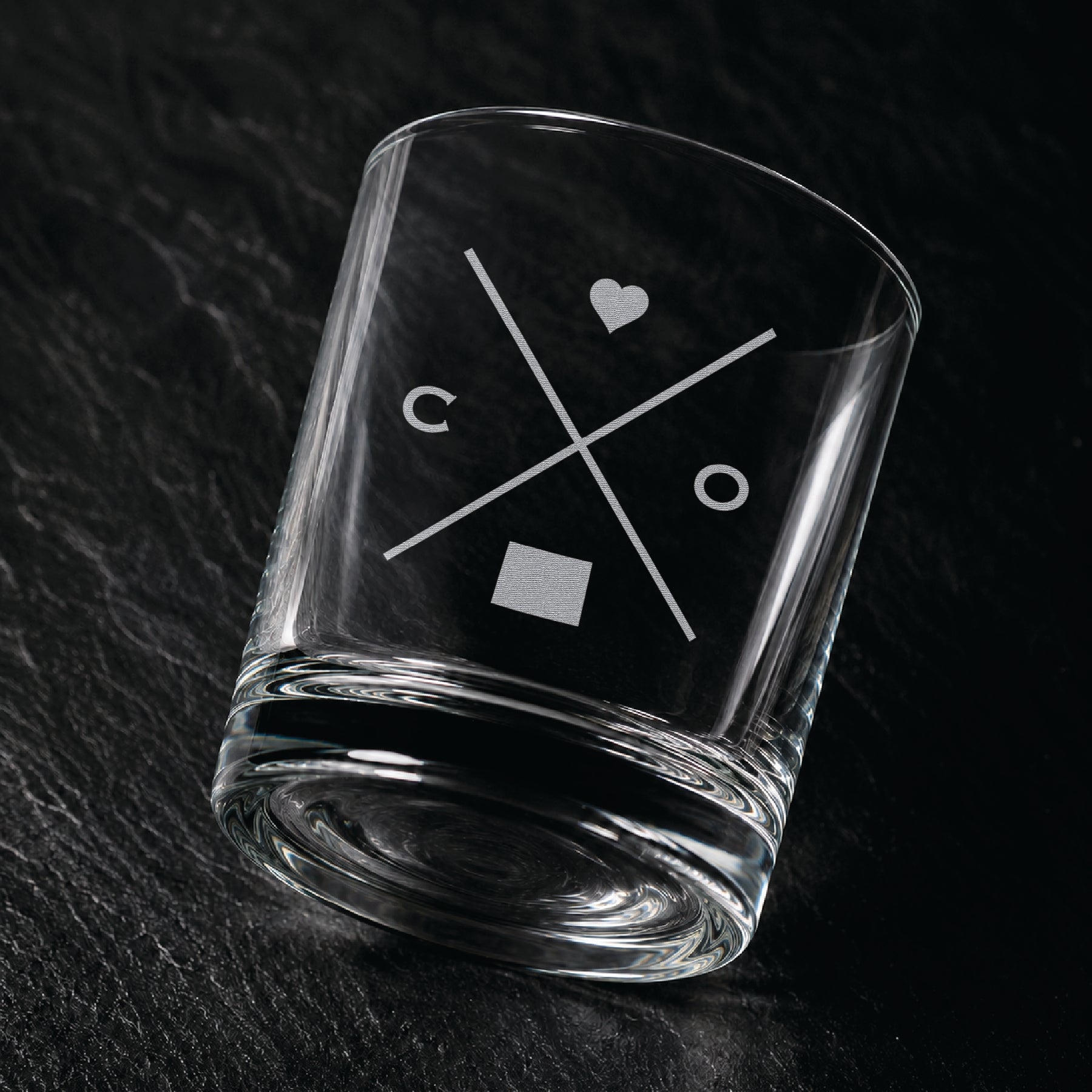 Angled engraved Colorado whiskey glass featuring frosted Colorado silhouette and modern crossed-letter design on a dark textured surface.