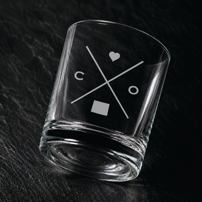 Angled engraved Colorado whiskey glass featuring frosted Colorado silhouette and modern crossed-letter design on a dark textured surface.