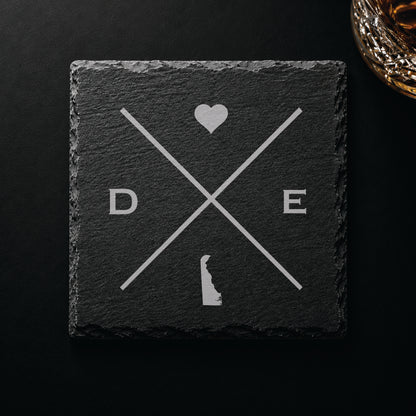 Dark slate coaster with engraved Delaware crossed-line design, heart icon, and Delaware silhouette on a dark background.