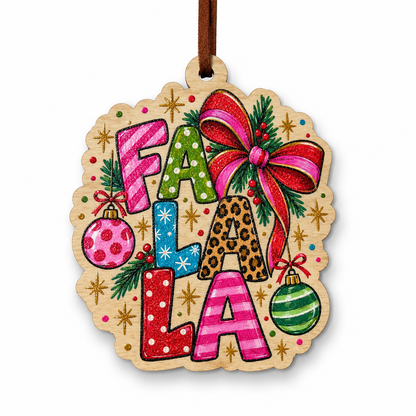 A festive wooden Christmas ornament featuring the words 'FA LA LA' in colorful patterned letters, decorated with a large red and pink bow, green pine branches, red berries, gold starbursts, and two hanging ornaments, suspended from a brown string on a white background.
