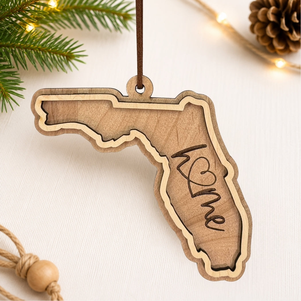 Layered Florida wood ornament featuring engraved “home” script and faux leather hanger on a soft neutral holiday background with greenery and warm lights.