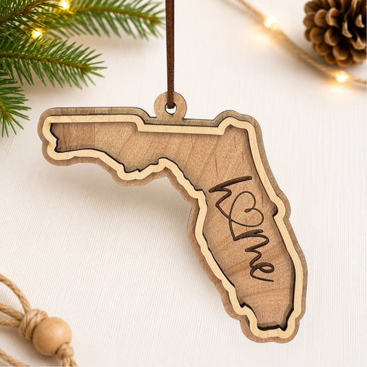 Layered Florida wood ornament featuring engraved “home” script and faux leather hanger on a soft neutral holiday background with greenery and warm lights.