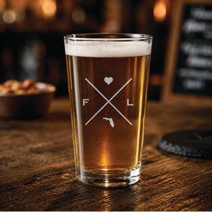 Engraved Florida pint glass filled with amber beer in a warm pub-style setting, featuring a minimalist crossed-line Florida design with heart detail.