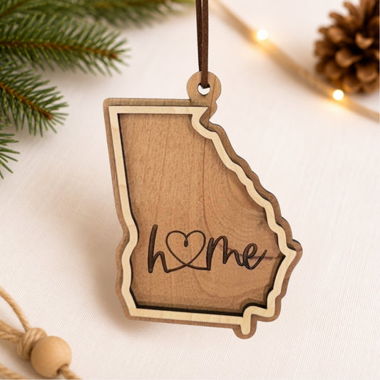 Layered Georgia wood ornament featuring engraved “home” script and faux leather hanger on a soft neutral holiday background with greenery and warm lights.