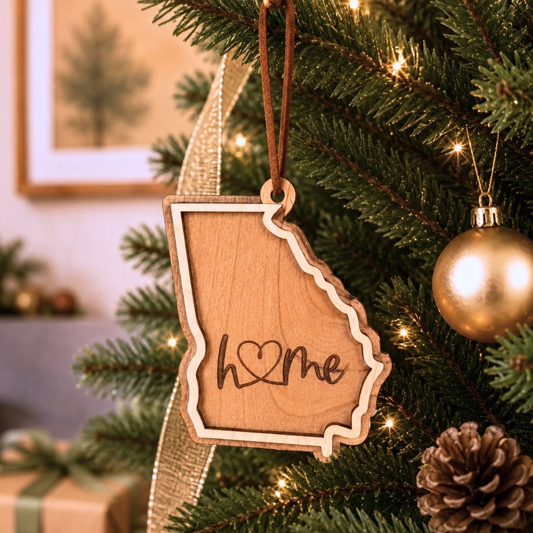 Layered Georgia “home” ornament hanging on a decorated Christmas tree with warm white lights, pinecones, and gold ornaments.
