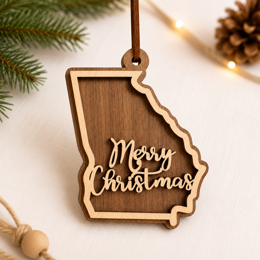 Layered hardwood Georgia Christmas ornament featuring walnut and maple wood with “Merry Christmas” script detail and faux leather hanger styled with evergreen branches and warm holiday lights.