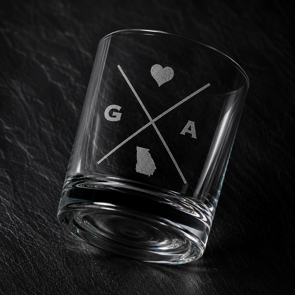 Angled engraved Georgia whiskey glass featuring frosted Georgia silhouette and modern crossed-letter design on a dark textured surface.