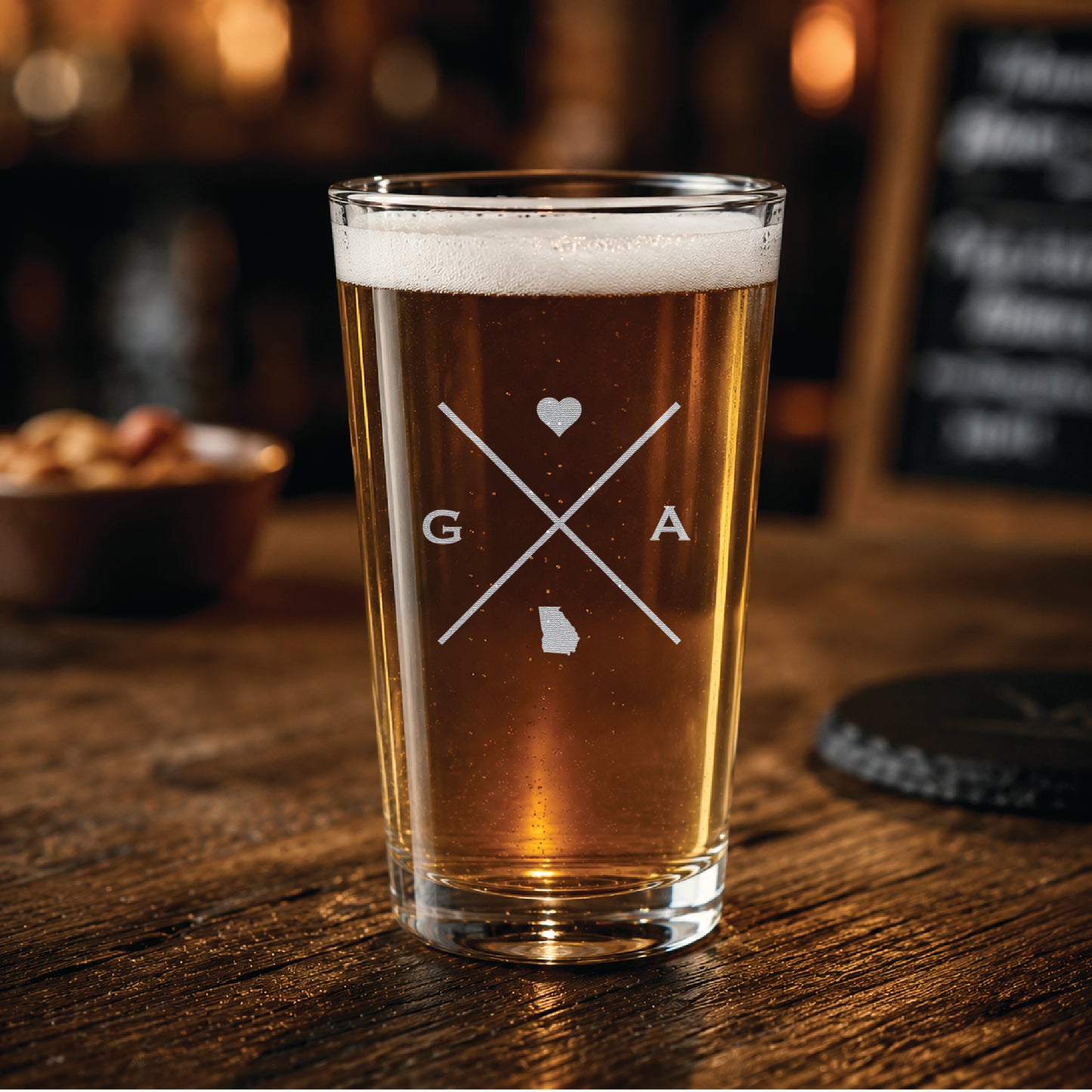 Engraved Georgia pint glass filled with amber beer in a warm pub-style setting, featuring a minimalist crossed-line Georgia design with heart detail.