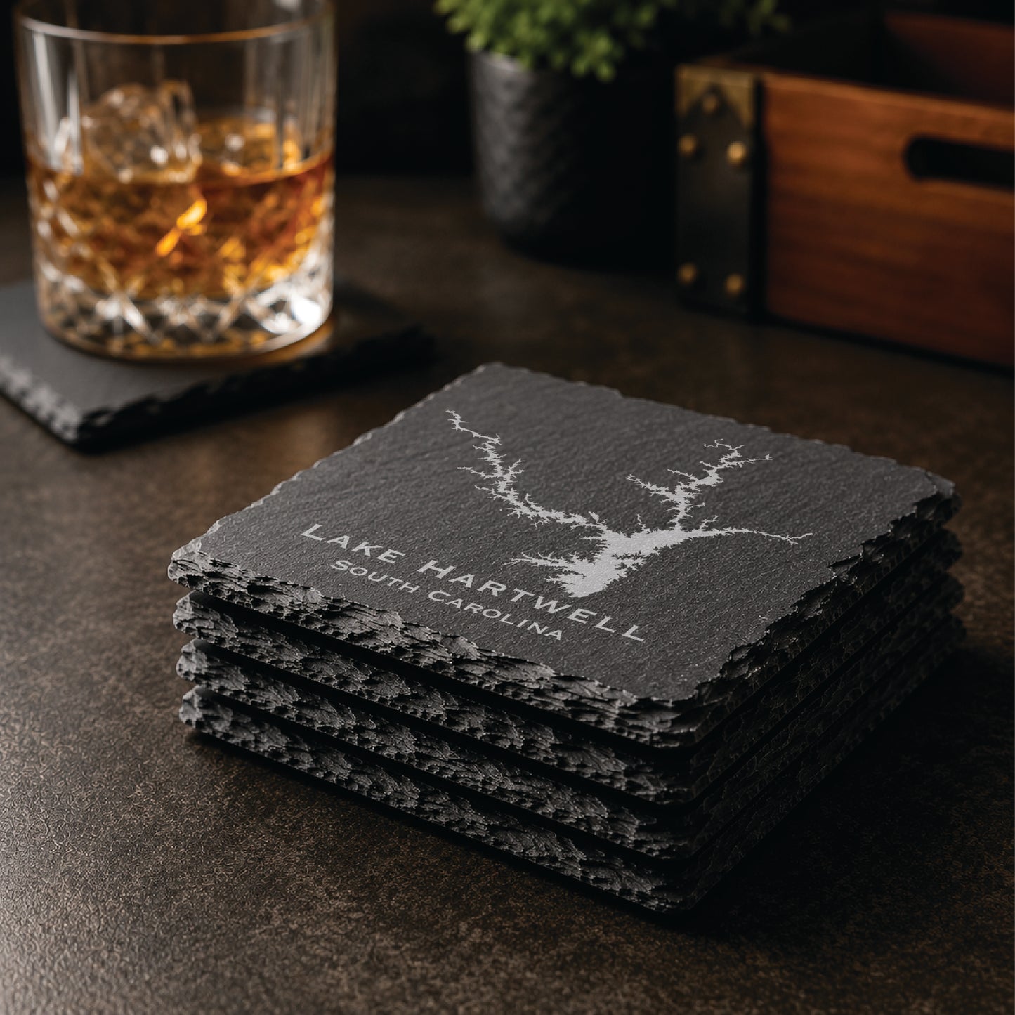 Stack of engraved Lake Hartwell slate coasters in a warm bar-style setting with whiskey glass nearby, featuring a detailed lake map design and South Carolina text.