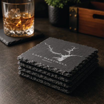 Stack of engraved Lake Hartwell slate coasters in a warm bar-style setting with whiskey glass nearby, featuring a detailed lake map design and Georgia text.