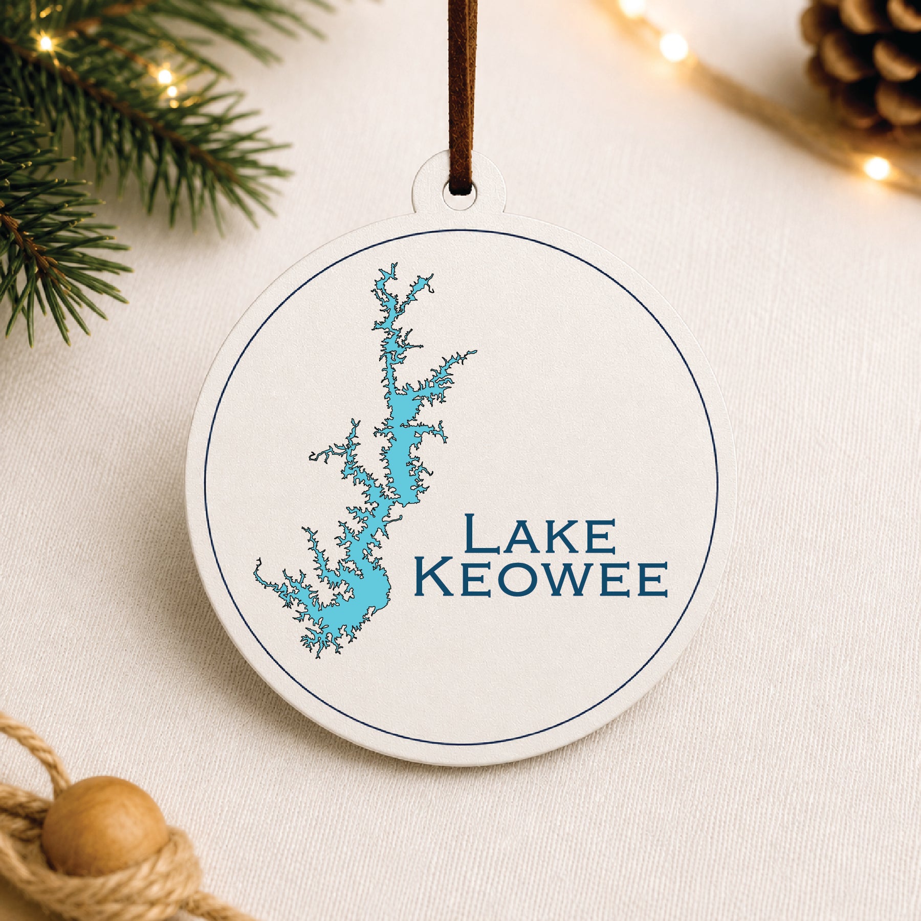 White Lake Keowee ornament with blue lake map design on a warm holiday linen background with greenery and lights.