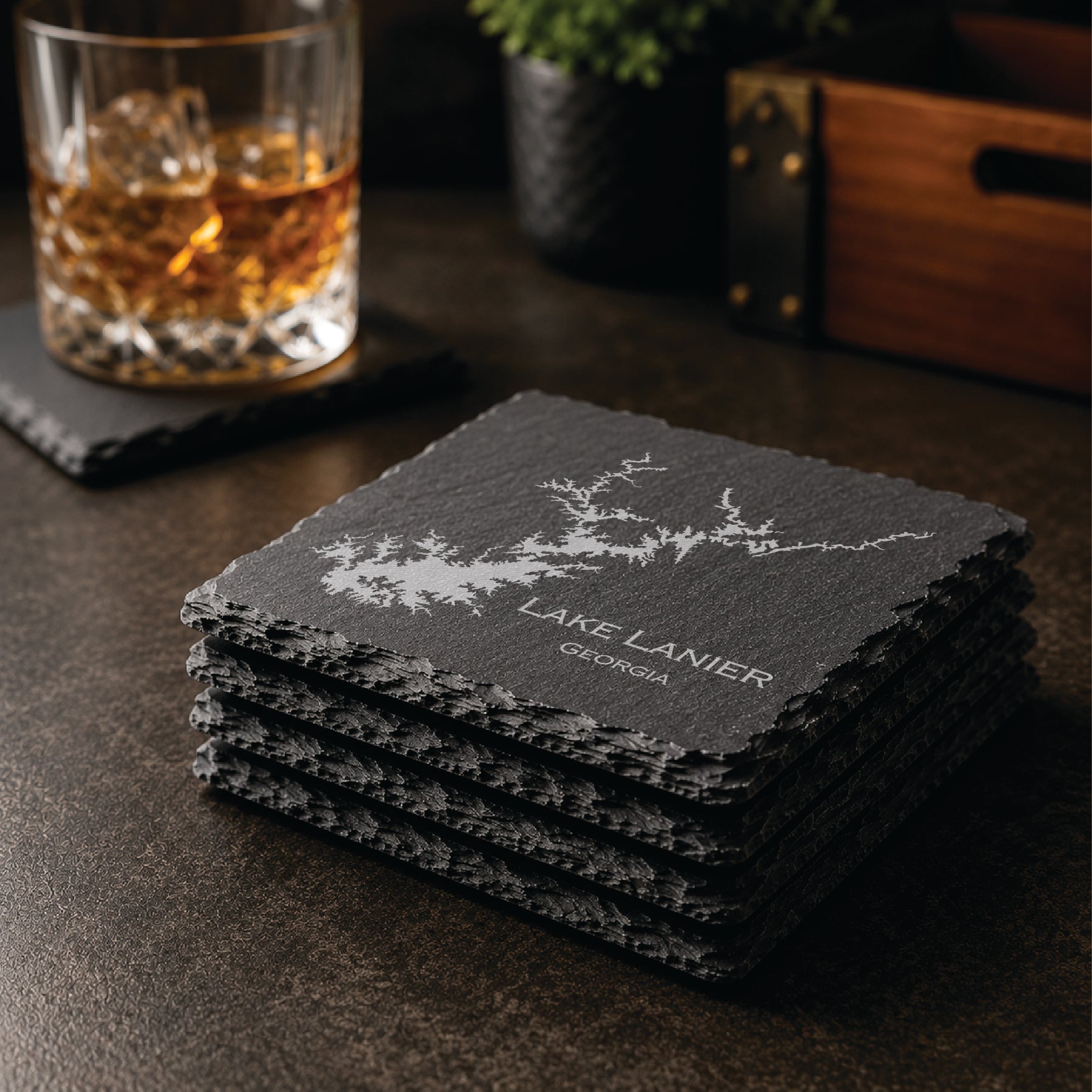 Stack of engraved Lake Lanier slate coasters in a warm bar-style setting with whiskey glass nearby, featuring a detailed lake map design and Georgia text.