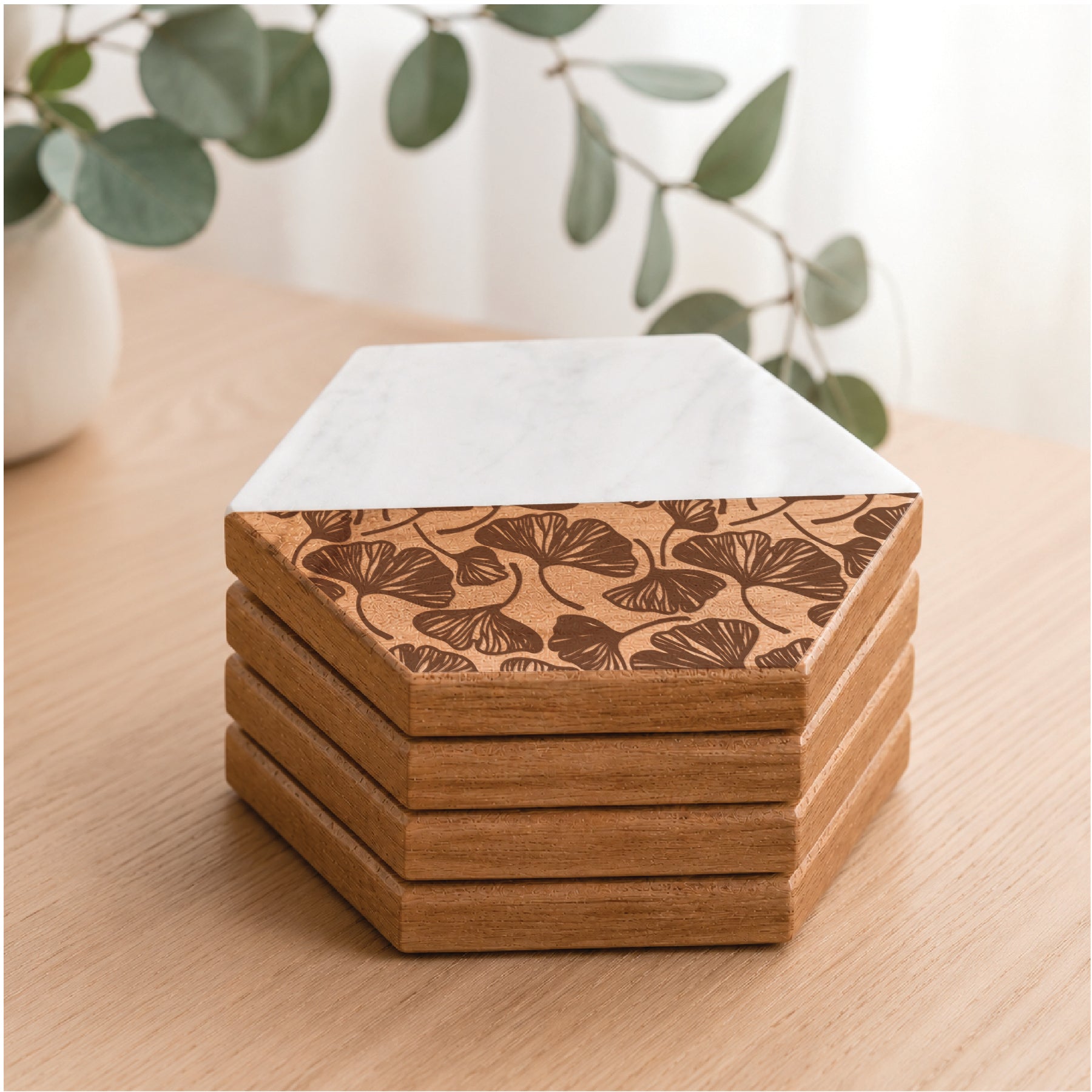 Stacked marble and acacia wood hexagon coasters with engraved botanical leaf pattern displayed on a light wood table with eucalyptus greenery in the background.