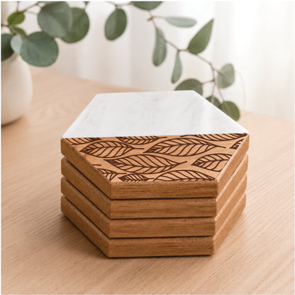 Stacked faux marble and acacia wood hexagon coasters with engraved falling leaves design displayed on a light wood tabletop with soft greenery in the background.
