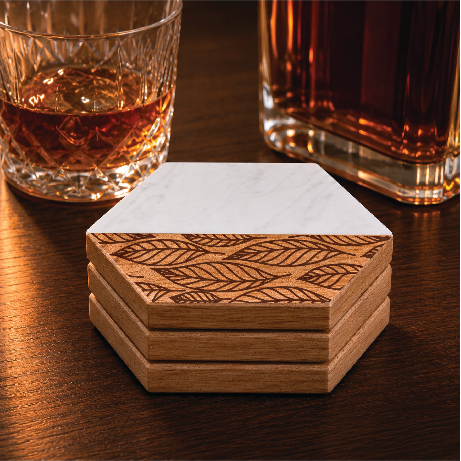 Set of four faux marble and acacia wood hexagon coasters featuring an engraved falling leaves pattern, styled beside a whiskey glass and decanter.
