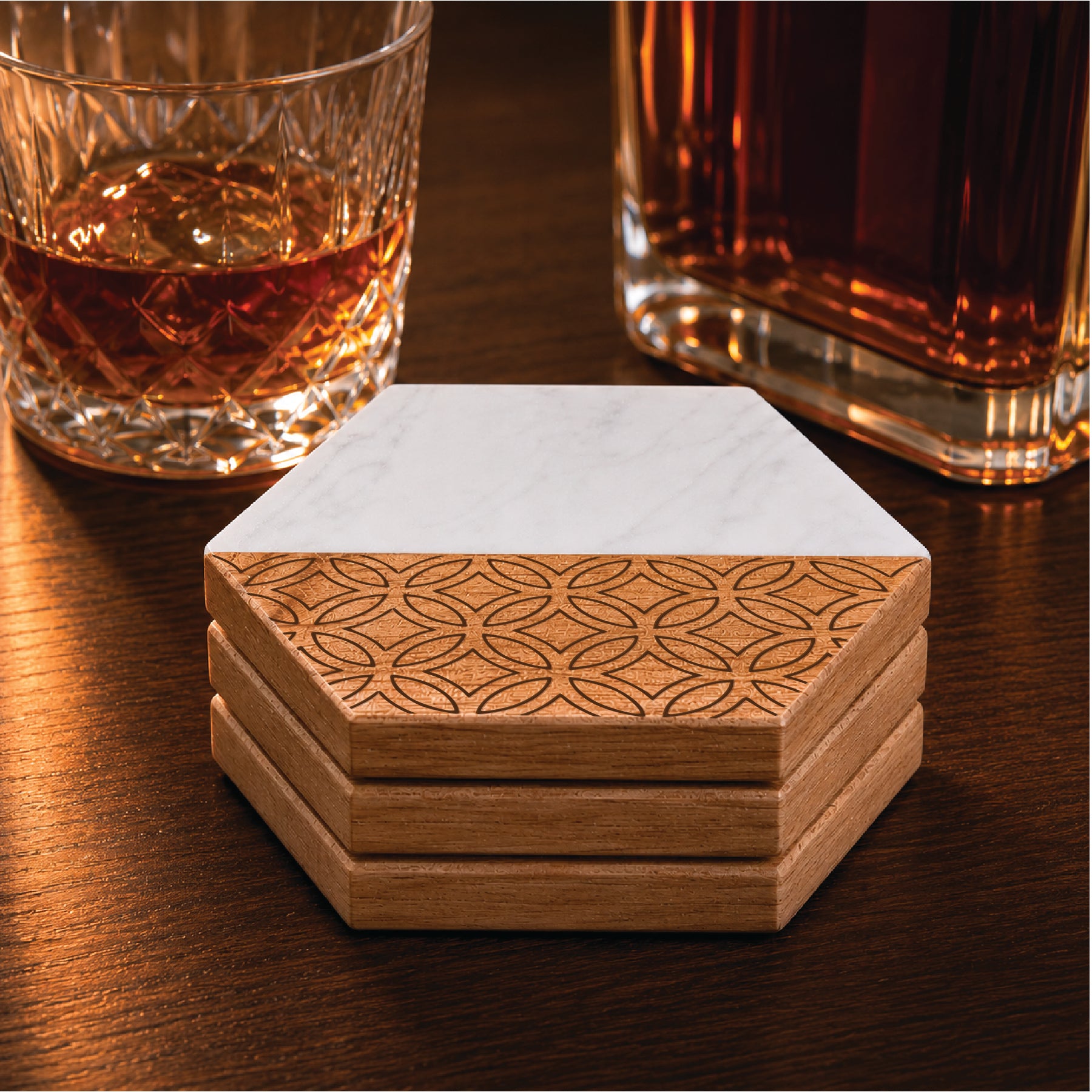 Stack of four hexagon marble and acacia wood coasters displayed beside a whiskey glass and decanter on a dark wood surface, highlighting the engraved geometric pattern and white marble design in a warm bar-inspired setting.