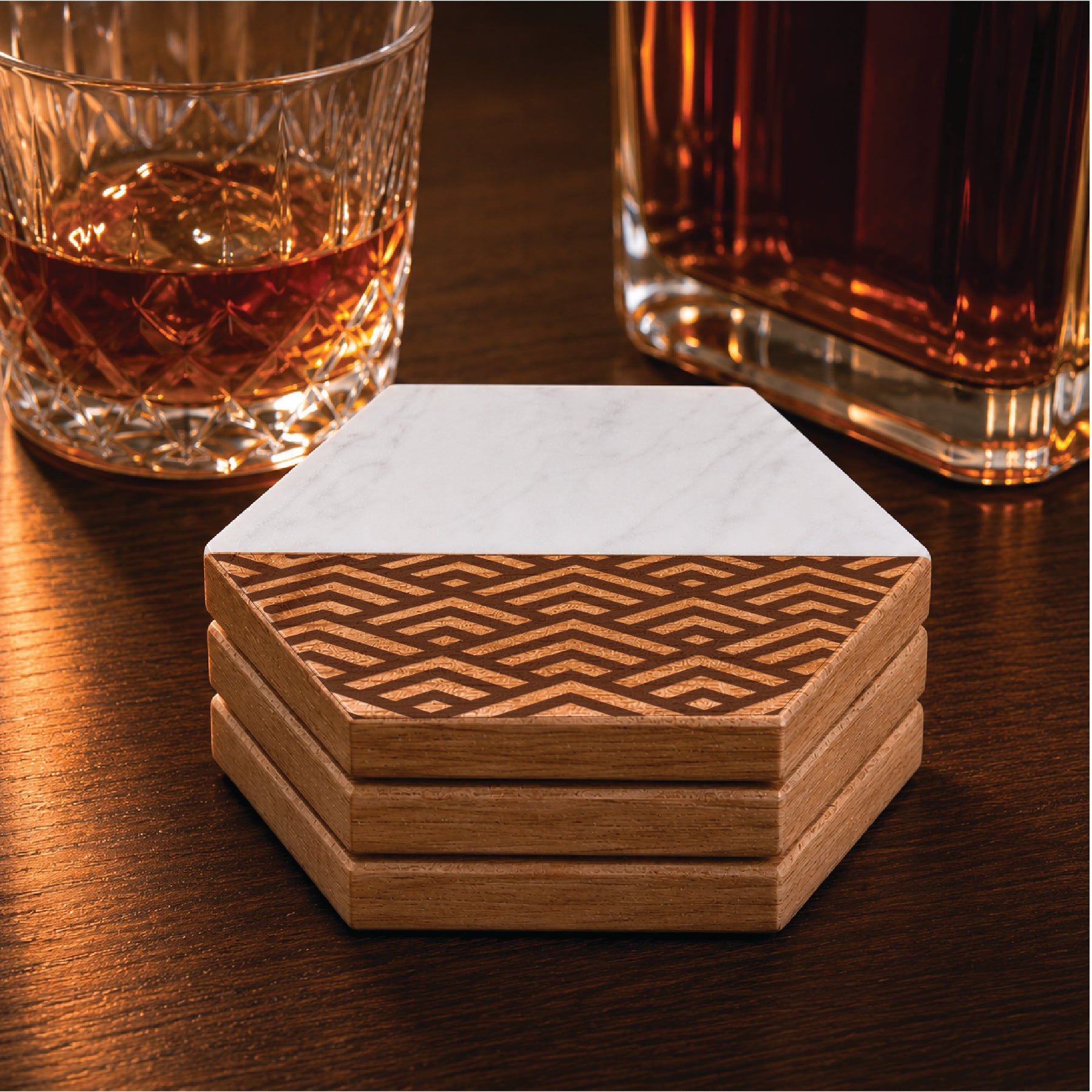 Set of four faux marble and acacia wood hexagon coasters featuring a geometric peaks pattern, styled beside a whiskey glass and decanter on a dark wood surface.