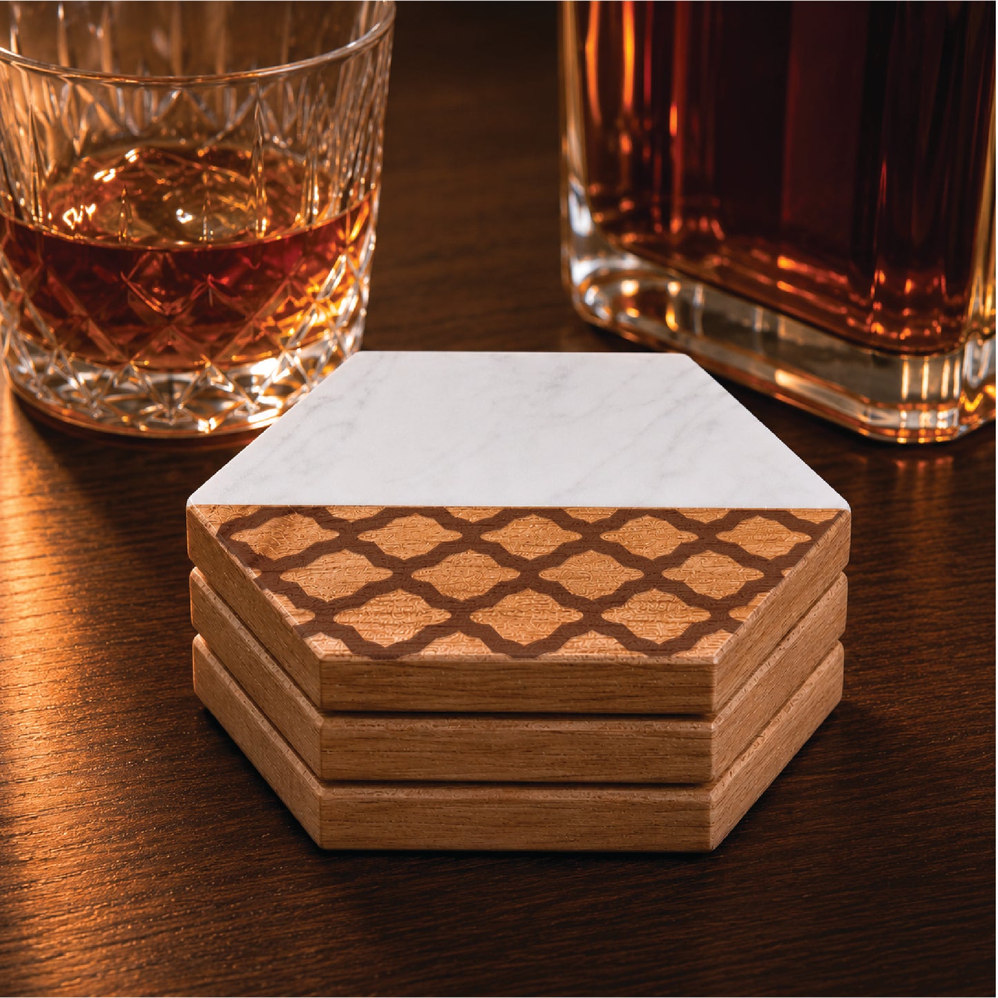Set of four faux marble and acacia wood hexagon coasters featuring a quatrefoil design, styled beside a whiskey glass and decanter on a dark wood surface.