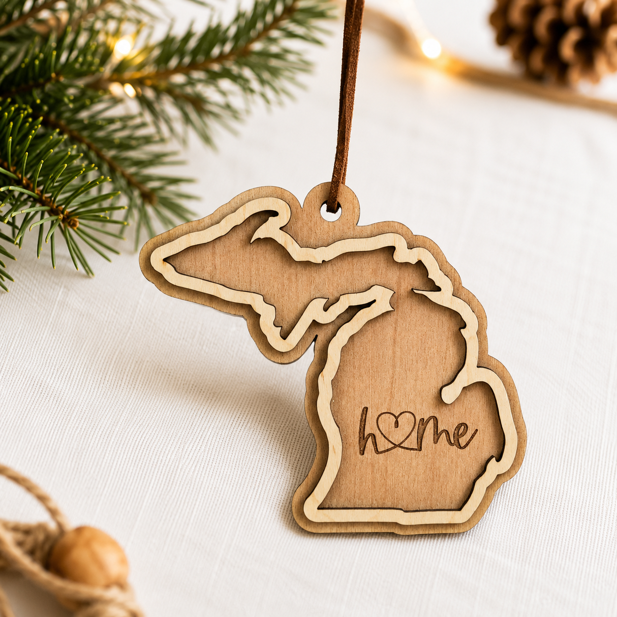 Layered Michigan wood ornament featuring engraved “home” script and faux leather hanger on a soft neutral holiday background with greenery and warm lights.