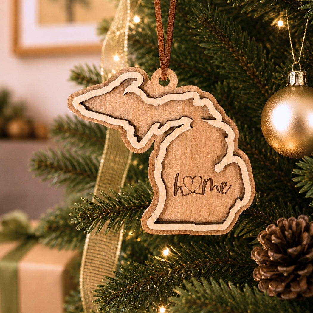 Layered Michigan “home” ornament hanging on a decorated Christmas tree with warm white lights, pinecones, and gold ornaments.