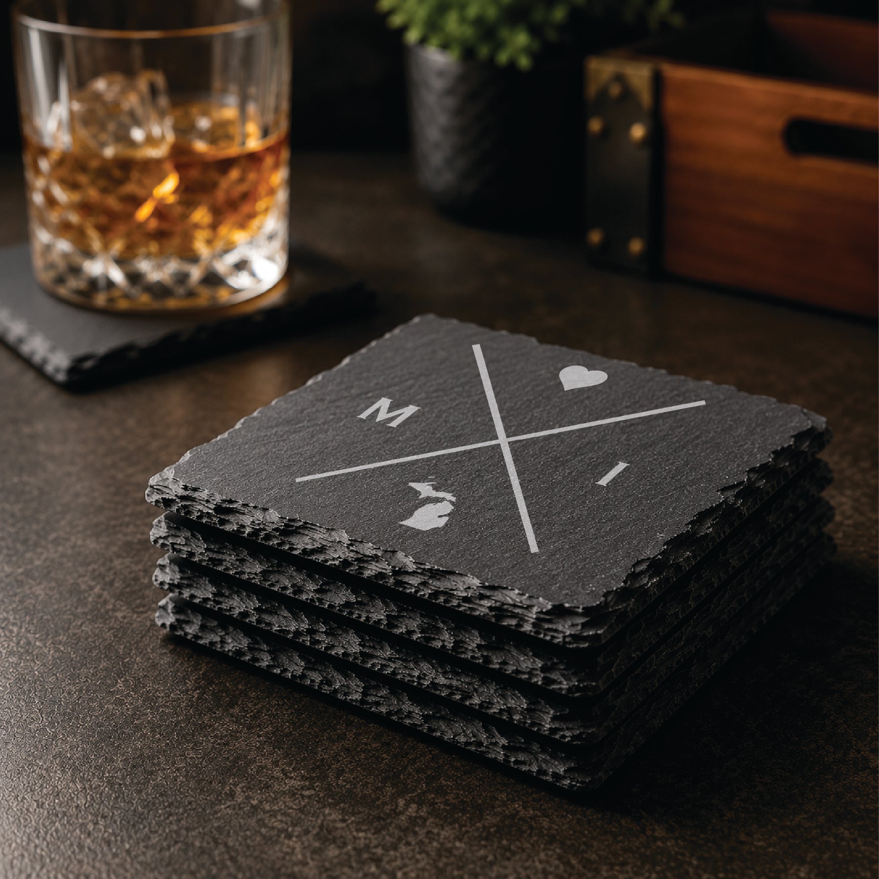 Stack of engraved Michigan slate coasters in a warm bar-style setting with whiskey glass nearby, featuring a minimalist crossed-line Michigan design.