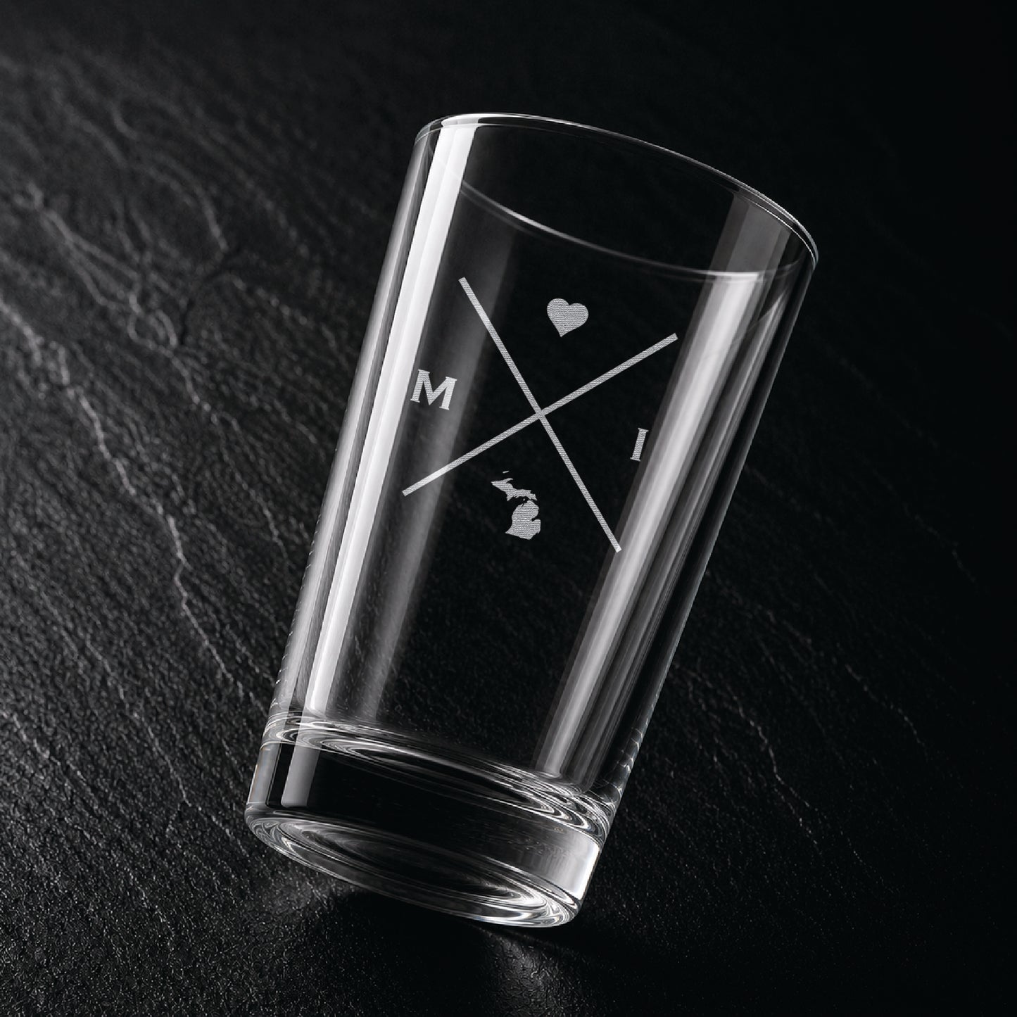 Angled view of engraved Michigan pint glass with modern crossed-line state design and frosted laser engraving on dark textured surface.