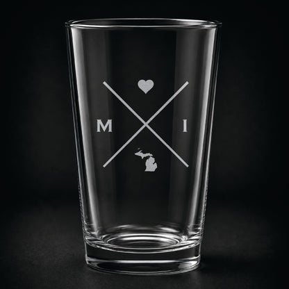 Engraved Michigan pint glass featuring a minimalist crossed-line design with heart detail and Michigan silhouette on a dark background.