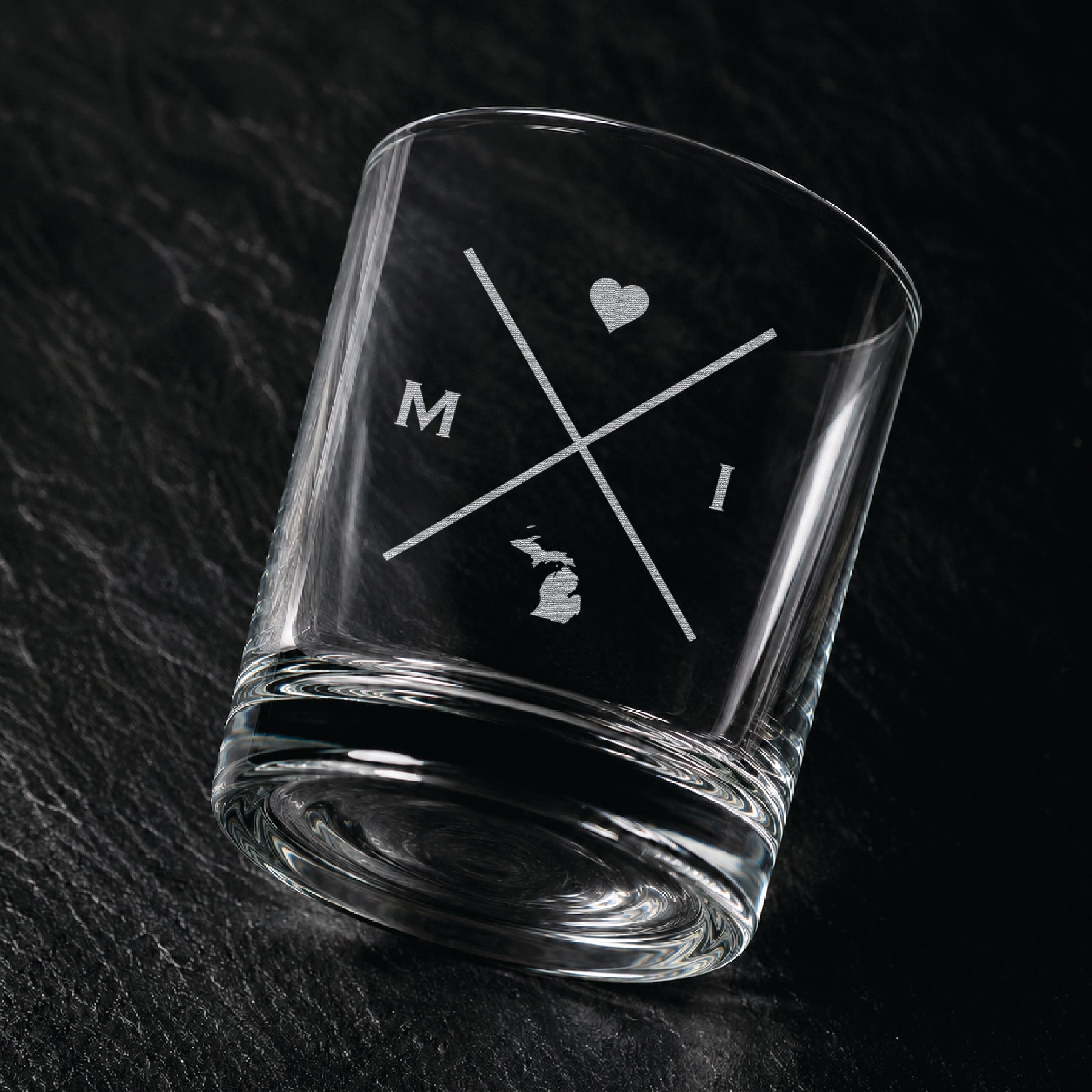 Angled engraved Michigan whiskey glass featuring frosted Michigan silhouette and modern crossed-letter design on a dark textured surface.