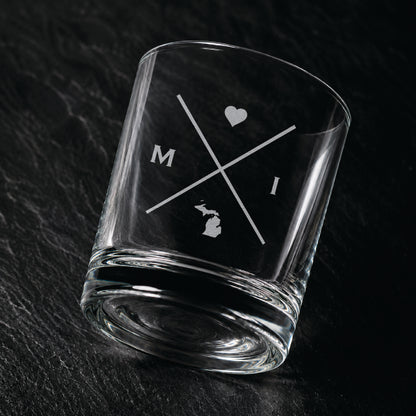 Angled engraved Michigan whiskey glass featuring frosted Michigan silhouette and modern crossed-letter design on a dark textured surface.
