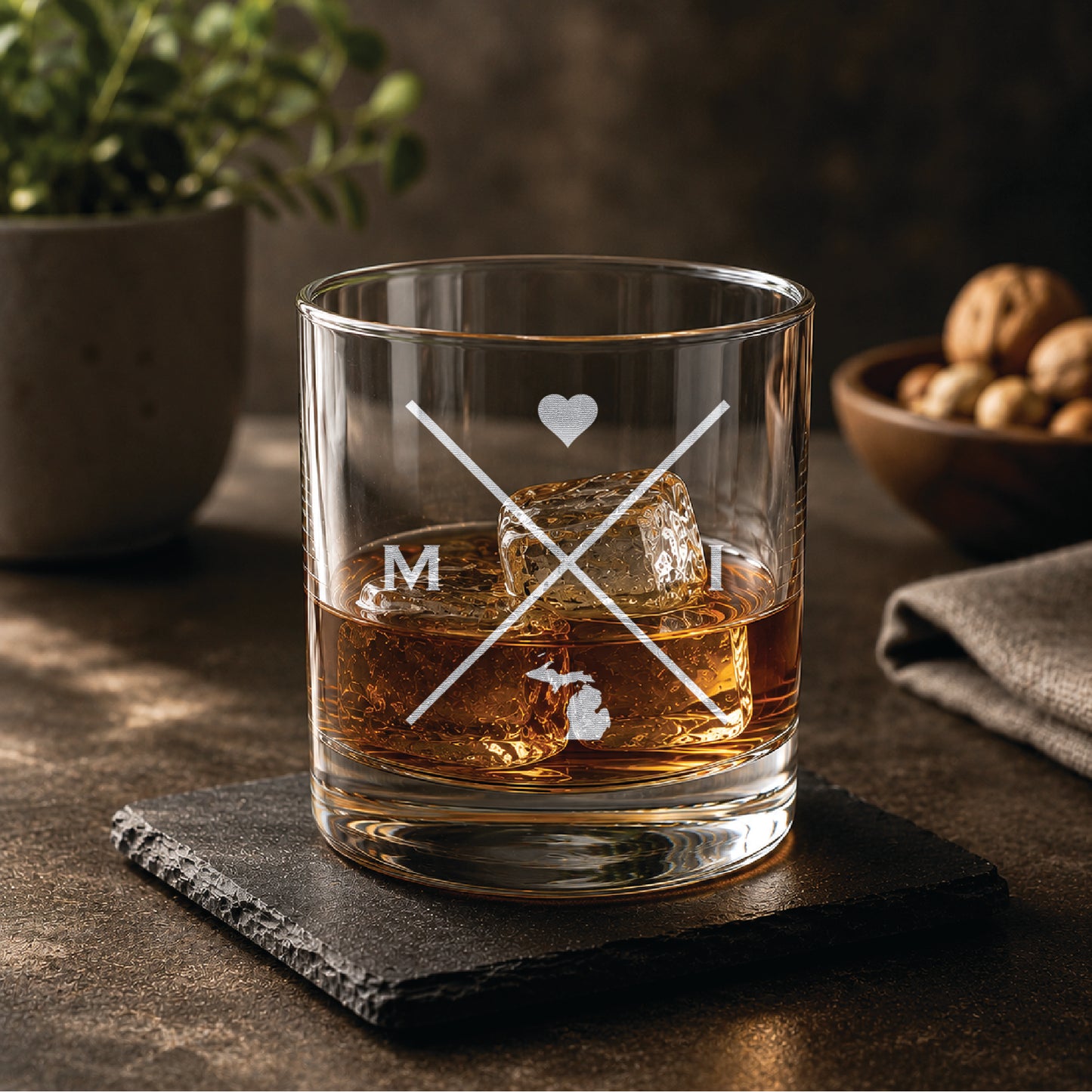 Engraved Michigan rocks glass with crossed-line monogram design, heart detail, and Michigan state silhouette, styled with whiskey and ice on a slate coaster in a warm, upscale bar setting.