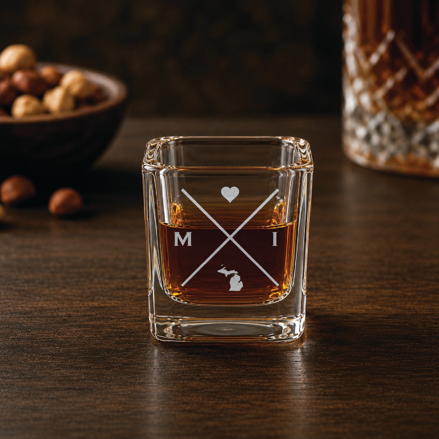 Engraved Michigan shot glass filled with whiskey in a warm bar-style setting, featuring a minimalist crossed-line design with heart detail and Michigan silhouette.