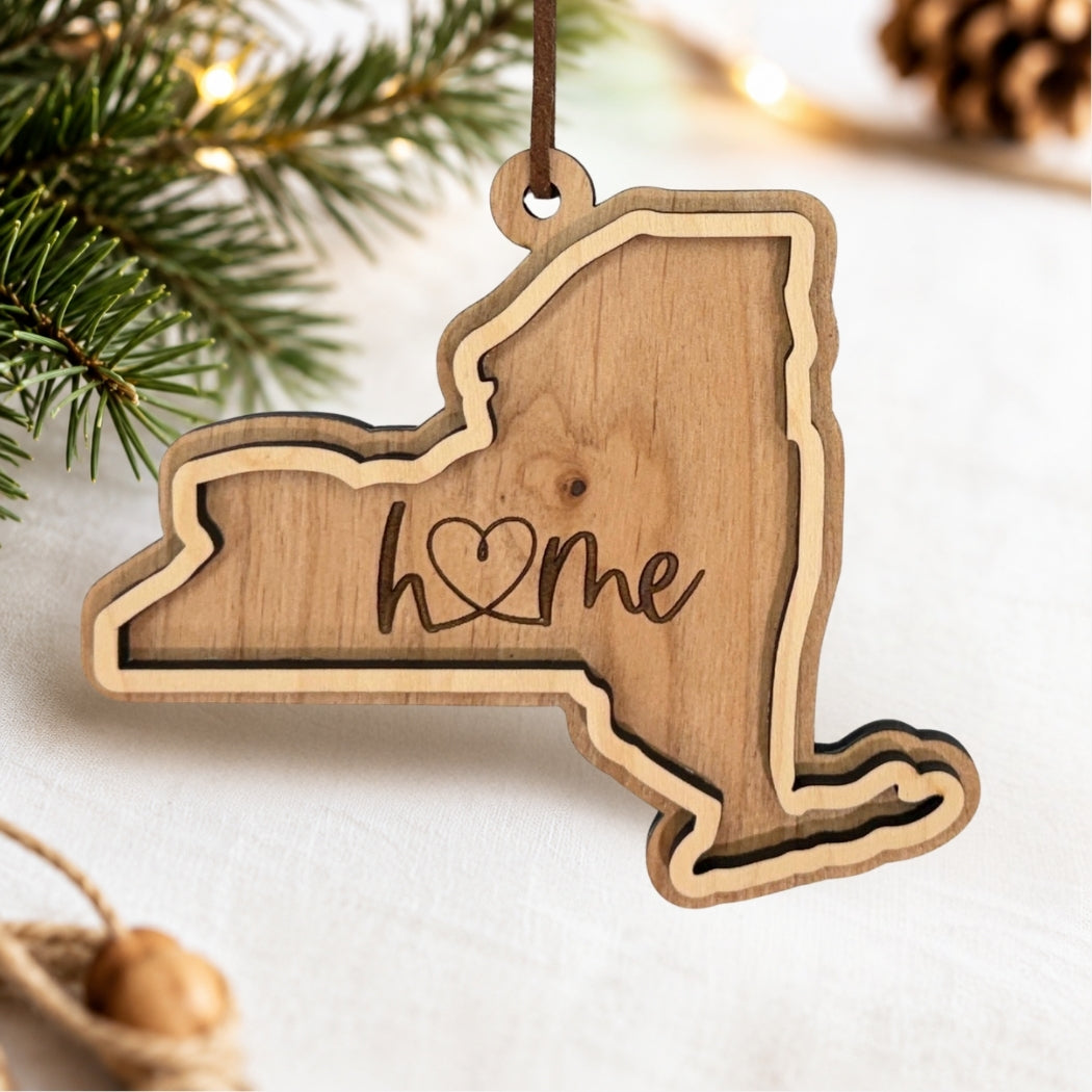 Layered New York wood ornament featuring engraved “home” script and faux leather hanger on a soft neutral holiday background with greenery and warm lights.